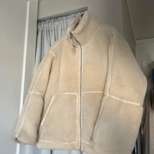 Beige teddyjacka - Mysig beige teddyjacka från H&M med hög krage och dragkedja framtill. Jackan har två fickor på sidorna och är tillverkad i ett fluffigt teddy-material som håller dig varm under kyliga dagar.