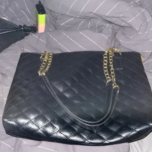 Svart quiltad axelväska med guldkedja - Snygg svart axelväska med quiltat mönster och guldfärgade kedjor som axelremmar. Väskan har dragkedja upptill och är rymlig med svart innerfoder. Perfekt för att lyfta din outfit med en elegant touch.