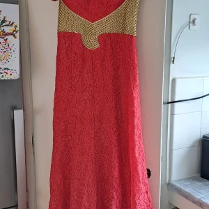Rosa långklänning med spets och gulddetaljer - Säljer en rosa långklänning med spets och dekorativa gulddetaljer över bysten och axelbanden. Klänningen har en stor rosett och tyllsläp baktill samt transparent mesh upptill. Perfekt för bal eller fest! Finns tillhörande bolero. Storlek är xs/s. Färgen är mer korall/rosa 