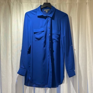 Blå skjorta från Primark - Säljer en blå skjorta med chiffong material från Primark med klassisk krage och två bröstfickor. Skjortan har långa ärmar med silvriga knappar vid manschetterna och framtill. 