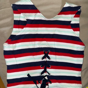 Randig klänning med snörning i ryggen - Säljer en färgglad randig klänning med marinblå, röda och vita ränder. Klänningen är ärmlös och har snygg snörning i ryggen som detalj. Perfekt för sommarens alla tillfällen!