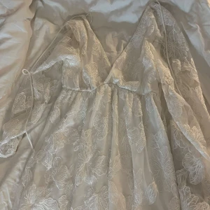Vit långklänning med blommigt mönster från Gina Tricot - Romantisk vit långklänning från Gina Tricot med v-ringning och transparenta, vida ärmar. Klänningen har ett vackert blommigt mönster i spets och markerad midja. Perfekt för sommarens speciella tillfällen. ANVÄND ENDAST EN GÅNG! Nypris: 800