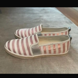 Randiga slip-on skor i rosa och vitt - Slip-on skor från GUESS med breda rosa och vita ränder. Skorna har platt sula. Perfekta för en avslappnad stil och passar bra till vår och sommar. Helt nya. 