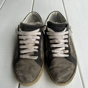 Saint Laurent grå sneakers med svart detalj - Grå sneakers från Saint Laurent med svarta detaljer och vit sula. Sidan har handskriven Saint Laurent-logga. Skorna har vita snören och är tillverkade i canvas och mocka. Box, påse och extra snören ingår