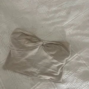 Beige ribbad bandeau tubtopp - Säljer en beige ribbad bandeau tubtopp med twistad detalj framtill. Toppen är ärmlös och har en mjuk, stretchig känsla som sitter snyggt på kroppen. Perfekt för varma dagar eller när du vill ha en enkel men trendig look.
