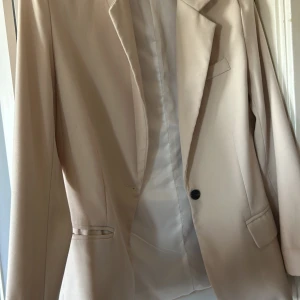 Beige kavaj med klassisk passform - Stilren beige kavaj med klassisk krage och en knapp framtill. Kavajen har två fickor med lock och en bröstficka. Fodrad insida och rak, tidlös siluett som passar till många olika looks. Perfekt för dig som gillar en clean och enkel stil. Från boohoo 
