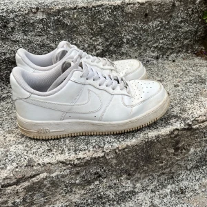 Nike Air Force 1 vita sneakers - Klassiska Nike Air Force 1 sneakers i helvitt skinn med perforerad tå, rund tå och platt sula. Snygg och tidlös modell med snörning och diskret Nike-logga på sidan och hälen. Perfekta för dig som gillar streetstyle och vill ha en ikonisk sneaker i garderoben.