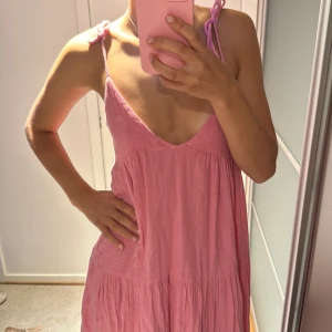 Rosa långklänning med knytaxelband - Säljer en luftig rosa långklänning med smala axelband som knyts upptill. Klänningen har djup v-ringning och volangig, flowy kjol. Perfekt för varma dagar och ger en chill vibe. Materialet känns lätt och skönt mot huden.