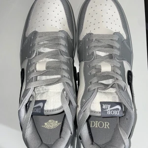 Nike Air Jordan 1 x Dior dunk sneakers - Exklusiva Nike Air Jordan 1 x Dior sneakers i vitt och grått skinn med grå snörning och ikonisk Dior-mönstrad svart swoosh. Tydliga Dior-detaljer på plösen, innersulan och genomskinlig yttersula med logga. Perfekt för dig som vill sticka ut med lyxig streetstyle. Pris kan diskuteras 