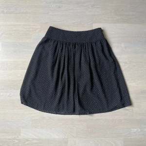 Svart prickig kjol från Burberry - Supersöt svart kjol från Burberry med vita prickar och bred midja. Kjolen är kort och har ett lätt utställt fall som ger en skön och luftig känsla. Perfekt för dig som gillar klassisk stil med en twist. Fodrad insida för extra komfort.