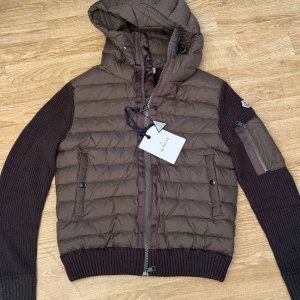 Moncler cardigan - Säljer en brun Moncler dunjacka i storlek M. Jackan har quiltad front, stickade ärmar och ribbade muddar. Utrustad med huva, dragkedja framtill och fickor med knappar samt en ärmficka med dragkedja. Perfekt för kyliga dagar och snygg till streetwear.