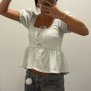 ljus grön blommig blus med puffärm - Supersöt ljus grön blus med små blommor, puffiga korta ärmar och knytning framtill. Modellen är lätt utsvängd nedtill och har en romantisk vibe. Perfekt för dig som gillar gulliga detaljer och en feminin look.