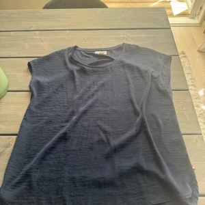 Mörkblå ärmlös linnetopp - En stilren mörkblå linnetopp med rund halsringning och avslappnad passform. Toppen är ärmlös och tillverkad i mjuk bomull, perfekt för varma dagar eller lager på lager. Enkel och clean design som funkar till många olika looks.