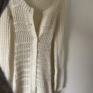 Strandklänning - Säljer en , vit stickad klänning med snyggt hålmönster och knappar hela vägen framtill. Klänningen har lång ärm och en klassisk rund halsringning. Perfekt att slänga över bikinin för en chill och stilren look.