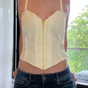 Gul korsett med spets och dragkedja - Söt ljusgul korsett med smala axelband, dekorativa spetsdetaljer och gul dragkedja framtill. Toppen har en feminin form med markerad midja och lätt v-formad nederkant. Perfekt för dig som gillar romantisk och trendig stil. Inga lappar på så vet ej vart ifrån den är!