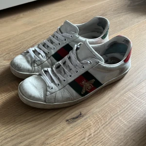 Gucci Ace  - Gucci Ace sneakers i vitt skinn med klassiska gröna och röda ränder på sidan samt broderad guldbi. Baksidan har metallicdetaljer i grönt och rött ormskinnsmönster. Platt sula och snörning framtill. Perfekta för dig som vill sticka ut med lyxig streetstyle. Priset kan diskuteras 
