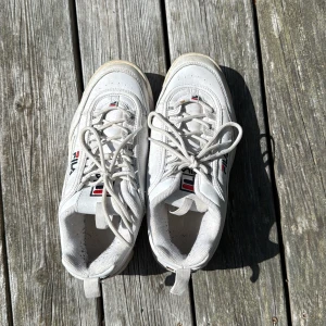 Vita chunky sneakers från FILA - Säljer ett par vita FILA Disruptor sneakers med chunky sula och klassisk logga på sidan, plösen och hälen. Skorna har snörning och är tillverkade i syntetmaterial och skinn. Perfekta för dig som gillar streetstyle och vill ha en sportig vibe.