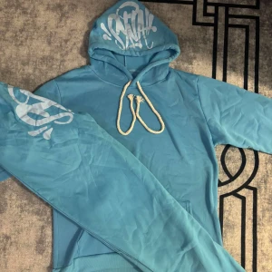 Syna World tracksuit Blå  - Säljer en turkos hoodie med vit graffiti-inspirerat tryck på huvan och ena ärmen. Tröjan har en stor känguruficka framtill och grova rep som snörning i huvan. Perfekt för dig som gillar streetwear och vill sticka ut med en färgglad look.