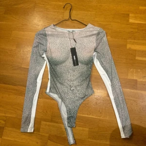 Vit och grå långärmad bodytop - Säljer en snygg långärmad bodytop  i vit och grått med småprickigt mönster. Toppen har figurnära passform, rund halsringning och vita paneler längs sidorna. Perfekt att styla med jeans eller kjol för en trendig look.