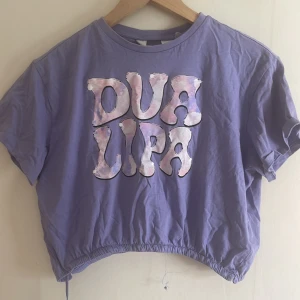 Lila Dua Lipa t-shirt från H&M - Säljer en lila t-shirt från H&M med stort Dua Lipa-tryck i pastellfärger på bröstet. Modellen är croppad med dragsko nertill och har korta ärmar. Perfekt för dig som vill ha en cool och avslappnad vibe.