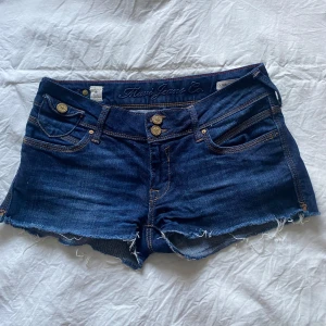 Mörkblå jeansshorts från Mavi midjemått 36 37  - Snygga mörkblå jeansshorts från Mavi med råa kanter och dubbla knappar framtill. Klassisk femficksmodell med coola detaljer och slitna benslut. Shortsen är tillverkade i bomull med lite stretch för skön passform. Perfekta för varma dagar och en avslappnad look.
