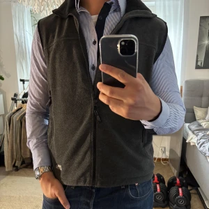 Svart fleeceväst från True North - Mysig svart fleeceväst från True North med hög krage och hel dragkedja framtill. Västen har två praktiska fickor med dragkedja och en stilren, enkel design. Perfekt att slänga på över en skjorta eller hoodie för extra värme.