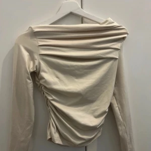 Beige omlottopp från SHEIN - Säljer en beige omlottopp från SHEIN i storlek S. Toppen har långa ärmar, draperad design och snygga rynkade detaljer på sidan. Materialet är mjukt och stretchigt, perfekt för en trendig look. Passformen är tight och axlarna är lätt avskurna för en cool siluett.