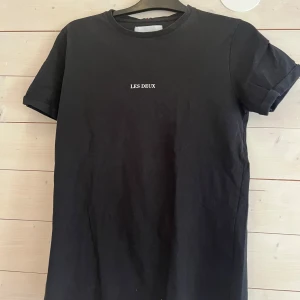 Svart t-shirt från Les Deux - Svart t-shirt från Les Deux med enkel vit text på bröstet. Klassisk rund halsringning och korta ärmar. T-shirten är i mjuk bomull och har en clean, stilren look som funkar till allt.