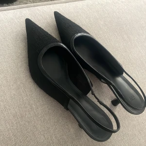 Svarta slingback klackar med spetsig tå från H&M - Enbart använda 1 gång, super trendiga & just dessa säljs ej längre i detta snygga materialet! Storlek 40 men passar 39❤️Stilrena svarta pumps från H&M med spetsig tå och låg klack. Skorna har en elegant slinga runt hälen och ovandel i textil med diskret struktur. Perfekta för dig som gillar en clean och trendig look.