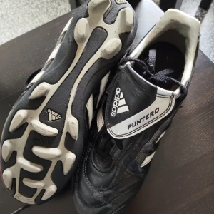 Adidas Puntero svarta fotbollsskor - Svarta Adidas Puntero fotbollsskor med vita detaljer och klassiska ränder på sidorna. Skorna har snörning och en robust sula med dobbar för bra grepp på planen. Tillverkade i syntetmaterial och skinn för slitstyrka och komfort. Storlek 36