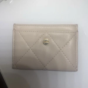 Beige quiltad plånbok i skinn - Snygg beige plånbok med quiltat mönster och guldfärgad knappdetalj. Slimmad design med flera fack för kort och sedlar. Perfekt att ha i fickan eller en mindre väska. Stilren och enkel att matcha till allt.