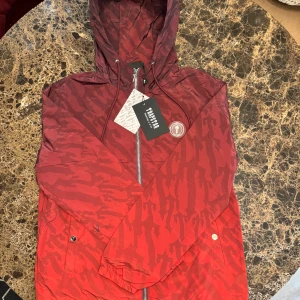 Röd camo vindjacka från Trapstar - Snygg röd vindjacka från Trapstar med camo-mönster och stor logga på ryggen. Jackan har huva med snörning, dragkedja framtill och två fickor med knapp. Perfekt för dig som vill sticka ut med streetwear-vibe.