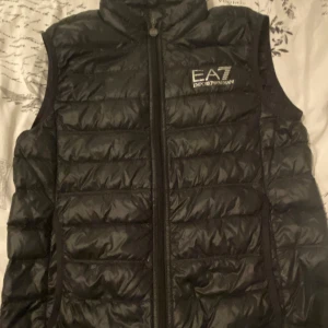 Svart dunväst från EA7 Emporio Armani - Snygg svart dunväst från EA7 Emporio Armani med quiltad design och hög krage. Västen har dragkedja framtill med EA7-logga på bröstet och på dragkedjans zip. Perfekt för lager-på-lager och ger en sportig vibe.