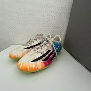 adidas fotbollsskor - Snygga fotbollsskor från adidas med vit bas och färgglada detaljer i blått, orange, rosa och gult. Skorna har svarta ränder på sidorna, snörning och en grön sula med röda dobbar. Perfekta för dig som vill sticka ut på planen.