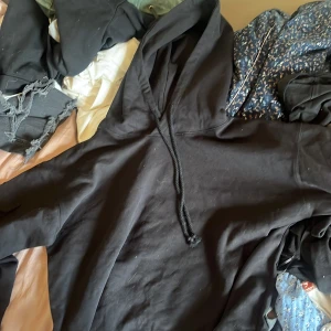 Svart hoodie med ficka på ärmen - Svart hoodie med huva och snörning, tillverkad i mjuk bomull. Tröjan har en unik ficka med lock på ena ärmen som ger en cool och praktisk detalj. Perfekt för dig som gillar streetwear och vill ha något extra i din garderob.