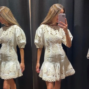 Vit kort klänning med broderade detaljer - Superfin vit kortklänning med puffärmar och broderade mönster. Klänningen har volang nertill och snygga utskurna detaljer som ger en unik look. Perfekt för sommarens alla tillfällen och ger en fräsch vibe.