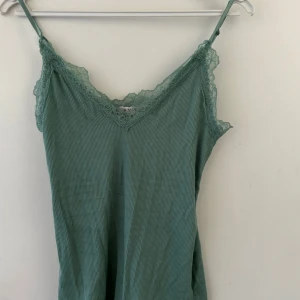 Grön ribbad linnetopp med spets - Snygg grön linnetopp med tunna axelband och v-ringning dekorerad med spets. Toppen är ribbad och har en mjuk, som passar perfekt till jeans eller kjol. Enkel men ändå detaljrik med spetsen som ger en extra touch.