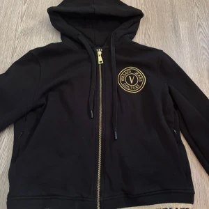 Svart hoodiejacka Versace Jeans Couture - Svart hoodiejacka från Versace Jeans Couture med guldfärgade detaljer och logga på bröstet. Som både passar S och XS. Jackan har dragkedja framtill, huva med snörning och ribbade muddar med guldfärgad text. Perfekt för en snygg och avslappnad streetstyle.