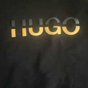 Svart sweatshirt från Hugo Boss - Svart sweatshirt från Hugo Boss med stort HUGO-tryck i gult och grått på bröstet. Klassisk rund halsringning och långa ärmar. Perfekt för dig som gillar stilrena och trendiga plagg med en tydlig logga framtill.