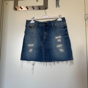 Blå jeanskjol med slitningar - Snygg blå jeanskjol med slitna detaljer framtill och fransig nederkant. Klassisk femficksmodell med knapp och dragkedja. Perfekt för dig som gillar en avslappnad och trendig stil. Från märket Odd Molly i storlek 1 som ungefär motsvarar en Storlek S. Mycket fint skick, jag hittar inga synlig anmärkningar eller defekter. 