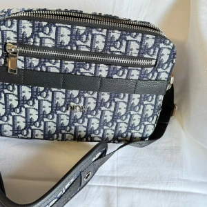 Dior axelväska i blått monogram - Säljer en snygg axelväska från Dior med klassiskt blått och vitt monogrammönster. Väskan har svarta skinn- och textildetaljer, silverfärgade dragkedjor och justerbar axelrem. Perfekt storlek för det viktigaste och riktigt trendig look.