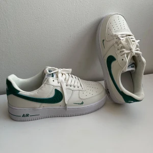Nike Air Force 1 vit/grön sneakers - Nike Air Force 1 sneakers i vitt skinn med gröna detaljer och swoosh-logga. Klassisk låg modell med perforerad tå, snörning och platt sula. Snygg kontrast mellan den vita basen och de gröna accenterna på häl och sidor.