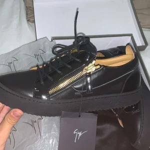 Svarta sneakers från Giuseppe Zanotti - Exklusiva svarta sneakers från Giuseppe Zanotti i blankt skinn med guldfärgade dragkedjor på sidorna och svarta snören. Snygg platt sula och beige innersula med guldlogga. Perfekt för dig som gillar statement-detaljer och lyxig känsla. Storlek 42