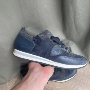 Philippe Model TRPZ skor  - Snygga sneakers från Philippe Model i mörkblått med camo-mönster. Skorna har detaljer i mocka och skinn, samt klassisk snörning. Sidan pryds av märkets logotyp och sulan är vit med svart yttersula. Perfekta för dig som gillar streetstyle med en twist. Nyskick. Storlek 41.