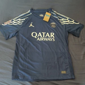 PSG x Jordan Dembélé #10 fotbollströja - Paris Saint-Germain x Jordan fotbollströja i mörkblått med vita detaljer och vingmönster på axlarna. Tryck med Qatar Airways, PSG-logga och Dembélé #10 på ryggen. Tillverkad i lätt och ventilerande polyester, perfekt för match eller träning.