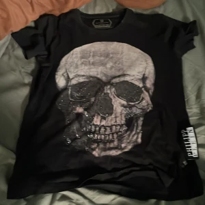 Svart t-shirt med dödskalle Philipp Plein - Svart t-shirt från Philipp Plein med stor dödskalle-tryck framtill och glittriga detaljer. Klassisk passform med korta ärmar och rund hals. Snygg metallogo bak i nacken och tydlig logotyp på sidan. Perfekt för dig som gillar edgy streetstyle.