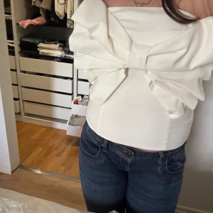 Vit axelbandslös topp med stor rosett - Superfin vit axelbandslös topp med en stor, iögonfallande rosett framtill. Sitter tight och har en elegant bandeau-form som ger en cool och trendig look. Perfekt för dig som vill sticka ut och älskar unika detaljer.