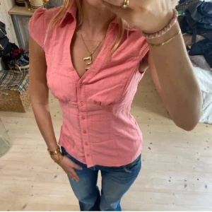 Rosa kortärmad skjorta med v-ringning - Snygg rosa skjorta med v-ringning och korta puffärmar. Skjortan har knappar framtill och figurnära passform som framhäver midjan. Perfekt till jeans för en fräsch och feminin look. Tunt, lätt material som känns skönt mot huden.