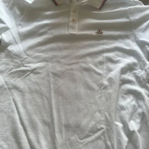 Vit pikétröja från Moncler - Snygg vit pikétröja från Moncler med klassisk krage och knäppning framtill. Tröjan har Moncler-logga broderad på bröstet och diskreta ränder i rött och marinblått på kragen. Tillverkad i 100% bomull för en skön och luftig känsla. Aldrig använd. Kom med förslag