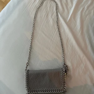 Grå axelväska med kedjedetaljer - Jag säljer denna väskan från Tiamo då den inte kommer till användning längre. Den är använd ett fåtal gånger och den är i ett bra skick. Kedjan har rostat lite precis vid början, men det är inget som syns.
